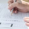 Close-up of a hand holding a pen and filling out printed forms on a desk. The forms have spaces for personal information such as name, address, and other details. The background is blurred, focusing on the hand and pen.