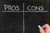 A blackboard is divided into two columns with the headings "PROS" and "CONS" written in white chalk at the top. A hand holding a piece of chalk is visible on the right side, preparing to write on the board.