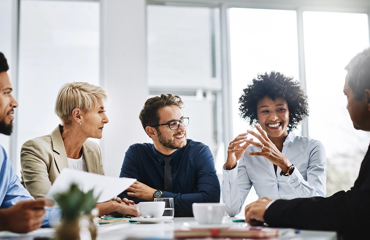 A group of five people sit around a table during a meeting in a bright office. They appear engaged in discussion, with one person gesturing while speaking. Papers and a tablet are on the table. Everyone is smiling and interacting positively.