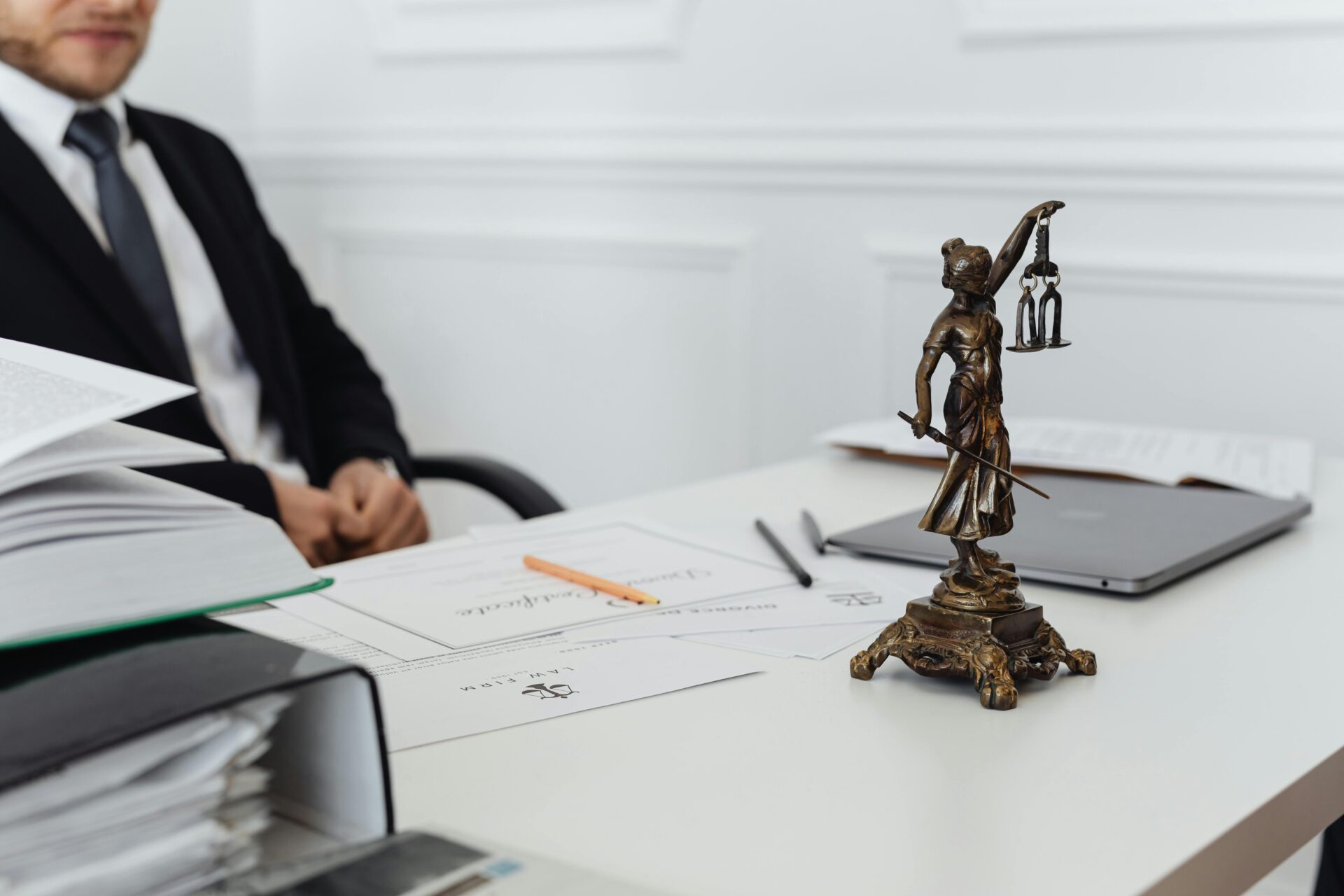 A person in a suit sits at a desk with documents, a laptop, and a pen, while a small bronze Lady Justice statue is prominently displayed in the foreground.