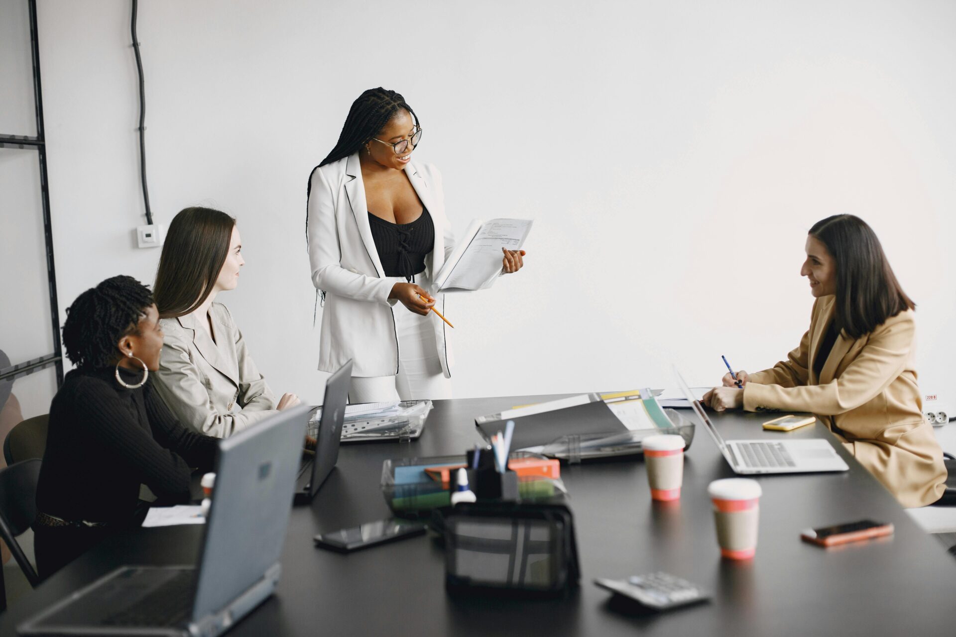 pexels-gustavo-fring-7446599 Four women are having a business meeting at a table with laptops and documents. One woman stands and presents, holding papers, while the others sit and listen attentively, taking notes.