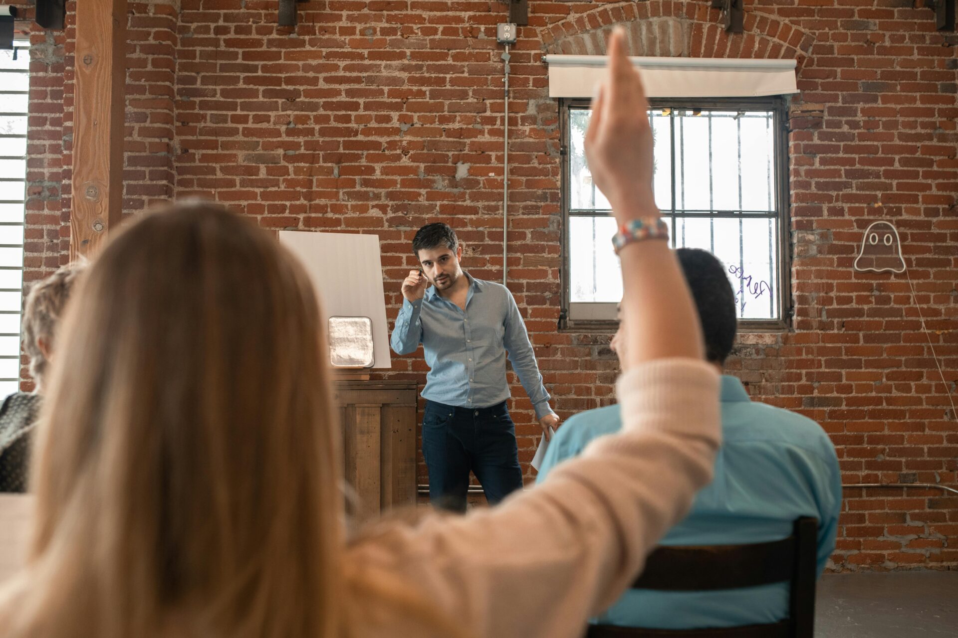 pexels-rdne-5756655 A man stands in front of a group, speaking and gesturing with his hand. A woman in the audience raises her hand to ask a question. The setting is a room with brick walls and large windows.