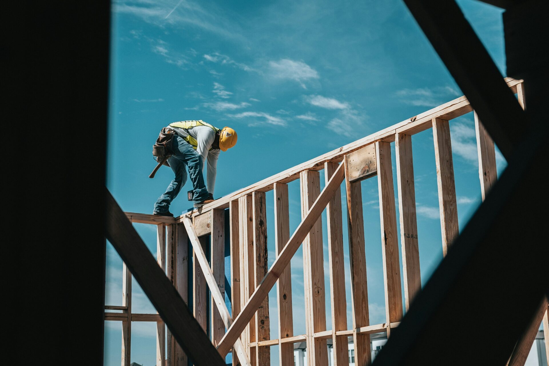 A construction worker wearing a yellow helmet and safety gear stands on a wooden frame of a building under construction, working under a clear blue sky.