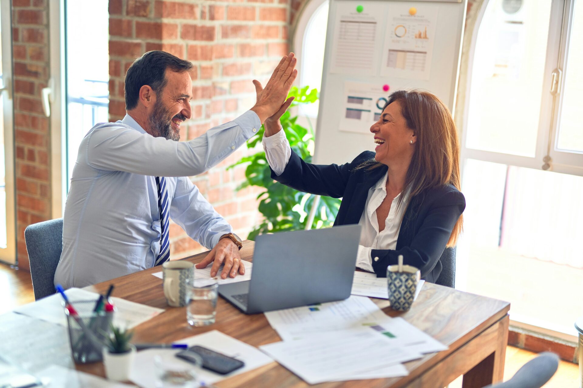 Two colleagues, a man and a woman, smile and give each other a high five while discussing Building Safer Workplaces: How Small Business Owners Can Strengthen Safety Policies at a desk with a laptop and documents in a bright office.