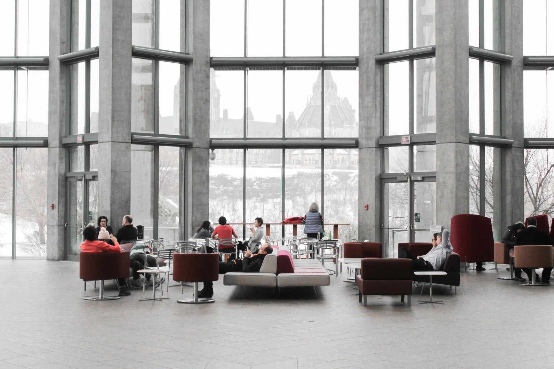 People sit and talk in a spacious, modern lobby with large windows overlooking a snowy landscape and historic building. Some discuss Compliance Traps for 25+ Employees, while others chat or relax on sofas and chairs.