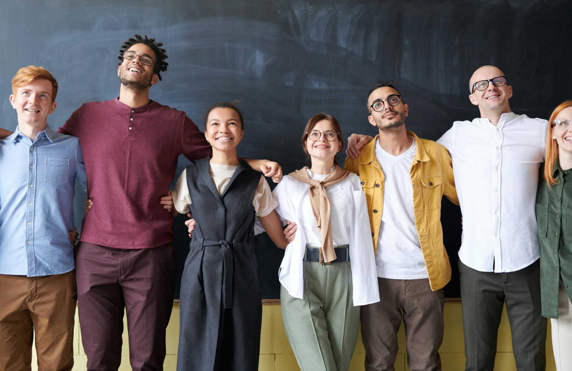 A diverse group of six people standing arm in arm, smiling and looking happy in front of a large blackboard. They appear to be friends or colleagues in a casual, friendly setting.