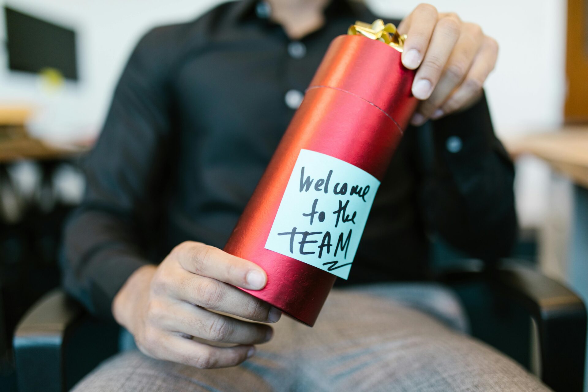 A person in business attire holds a red gift box with a note that says "Welcome to the TEAM" written in marker.