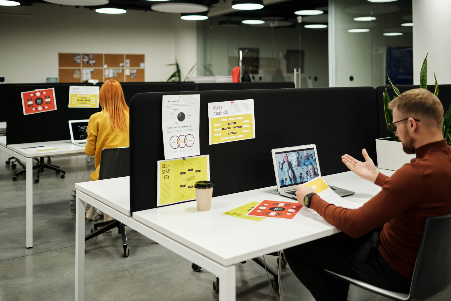 Two people work at separate desks in a modern office, divided by a black partition. One person participates in a video call on a laptop; papers, a coffee cup, and business charts are visible on the desks and partition.